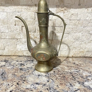 Puede incluir: Una tetera de latón con intrincados diseños florales y una tapa alta y puntiaguda. La tetera tiene un pico y una manija, y está decorada con acentos rojos, azules y dorados.