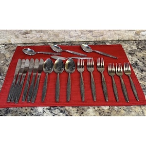 May include: A stainless steel flatware set consisting of six knives, three spoons, and six forks arranged on a red placemat. The set includes three additional spoons positioned horizontally at the top, all featuring a simple, polished metallic finish.