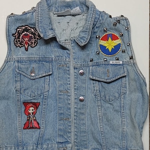 May include: Light blue denim vest with multiple character patches and silver stud accents. Patches include a red-haired character, a Captain Marvel logo, and a character in a red outfit. The vest has a button-up front and two front pockets.