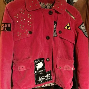 May include: A pink corduroy jacket with gold studs and various patches, including "Minor Threat", "The Adicts", and a white sheep. The jacket has a triangular patch with a symbol that resembles the Triforce from the Legend of Zelda video game series.