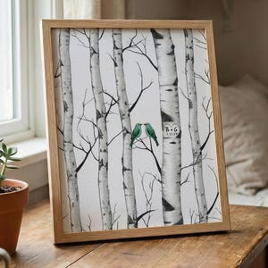 May include: A framed print depicting a black and white birch tree scene with two green birds. The print is in a light wood frame, placed on a wooden table. A small succulent in a terracotta pot and a coffee mug are also on the table.