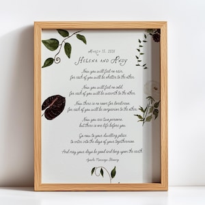 May include: A framed print with a light wood frame. The print features the names "Helena and Andy" and the date "March 15, 2026" in elegant script. The text is a marriage blessing, with botanical illustrations in green and burgundy.