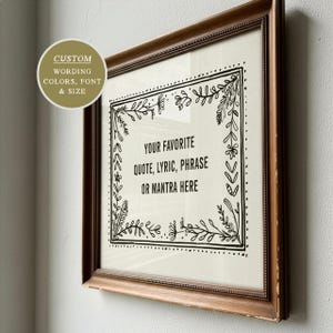 May include: A square wall art piece in a distressed brown wooden frame featuring a cream-colored background with a black ink botanical border. The center text reads: YOUR FAVORITE QUOTE, LYRIC, PHRASE OR MANTRA HERE. A circular overlay displays CUSTOM WORDING.