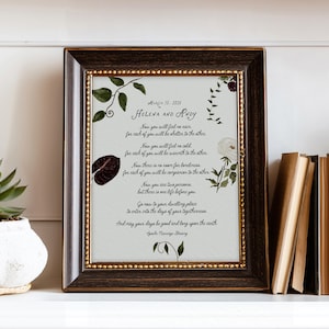 May include: A framed print featuring a wedding blessing, with the names "Helena and Andy" at the top. The text is surrounded by floral illustrations in green and burgundy. The frame is dark brown with a gold inner border. The print is displayed on a white shelf.