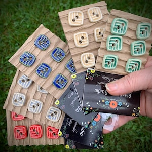 May include: Assortment of handmade ceramic buttons in various colors and patterns, including blue, green, yellow, and red. The buttons are displayed on brown cardboard cards with the text "Gift & Studio" and "Handmade Pottery" printed on them.