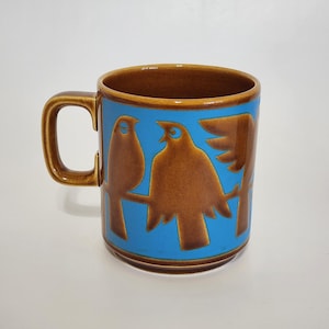 May include: A white ceramic mug with a black handle and the text "But First, Coffee" in a bold, black font. The mug is set against a blurred background, suggesting a cozy setting.