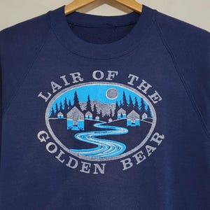 Puede incluir: Una sudadera azul marino de cuello redondo con un estampado gráfico dentro de un marco ovalado. El diseño muestra un río sinuoso, pinos y pequeñas cabañas bajo una luna, con el texto LAIR OF THE GOLDEN BEAR impreso en arco circular.
