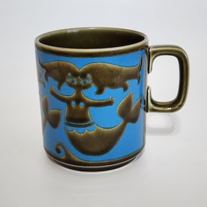 May include: A ceramic mug with a blue background and a brown mermaid design. The mug has a brown handle and rim. The mermaid has outstretched arms and a fishtail. The mug is a standard size for coffee or tea.