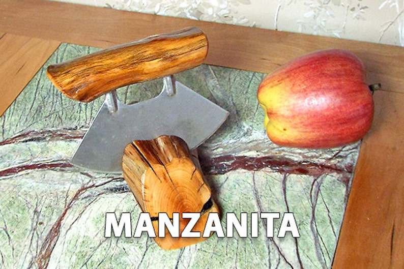 May include: A wooden apple corer with a stainless steel blade. The corer is made from manzanita wood and has a natural finish. The corer is sitting on a green and brown marble surface with a red apple next to it.