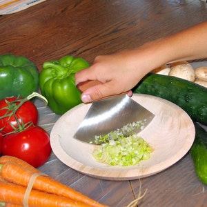 May include: A person is using a wooden handled, stainless steel chopping knife to chop green onions in a wooden bowl. There are green bell peppers, red tomatoes, carrots, cucumbers, and mushrooms on a white surface.