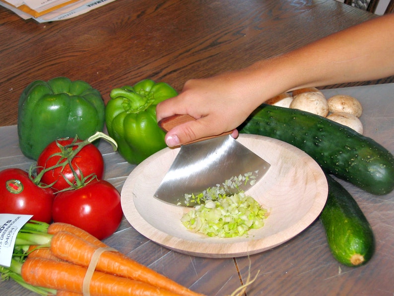 May include: A person is using a wooden handled, stainless steel chopping knife to chop green onions in a wooden bowl. There are green bell peppers, red tomatoes, carrots, cucumbers, and mushrooms on a white surface.