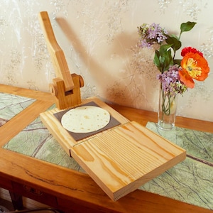 May include: A wooden tortilla press with a lever and a round, flat surface for pressing tortillas. The press is on a wooden table with a green marble inlay. A bouquet of purple and orange flowers is in a glass vase on the table.