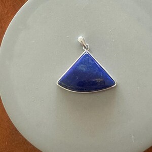 May include: A triangular pendant with a deep blue gemstone set in a silver-colored metal frame. The pendant is a vibrant blue with subtle variations in color. The bail is also silver-colored. The pendant is displayed on a light gray surface.