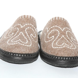 May include: A pair of brown felt slippers with white embroidered designs. The slippers have a black leather sole and a stitched upper. The design is symmetrical on each slipper, and the background is white.