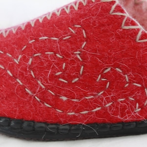 Pode incluir: Pantufla de feltro vermelho com um design branco costurado. A pantufla tem uma sola de borracha preta.