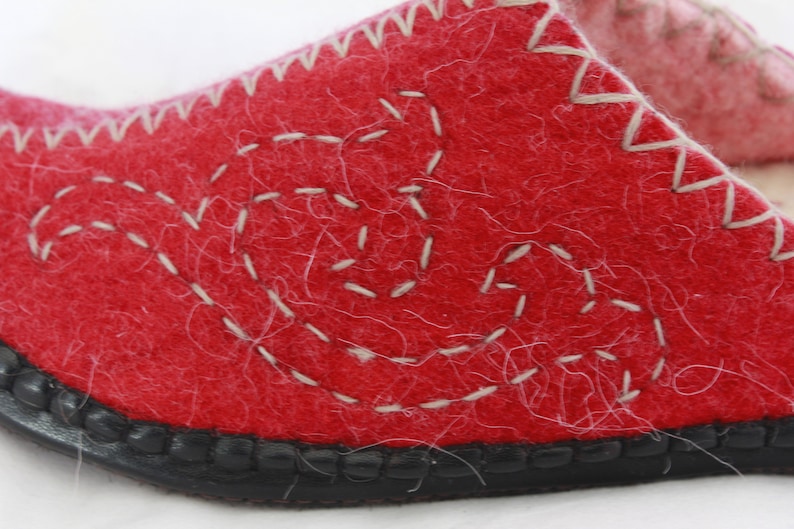 Pode incluir: Pantufla de feltro vermelho com um design branco costurado. A pantufla tem uma sola de borracha preta.