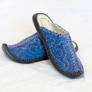 May include: A pair of blue felt slippers with white embroidered swirls and a black leather trim. The slippers have a soft, white interior and are designed for indoor wear. The stitching is a contrasting white color.