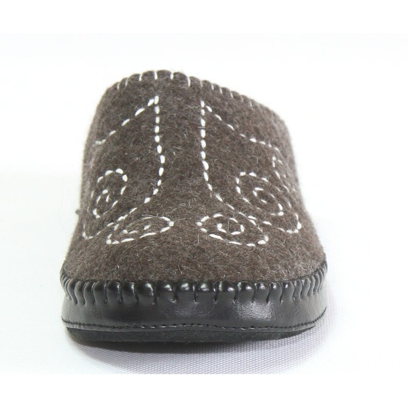 May include: A pair of brown felt slippers with white stitching in a decorative pattern. The slippers have a black leather sole.