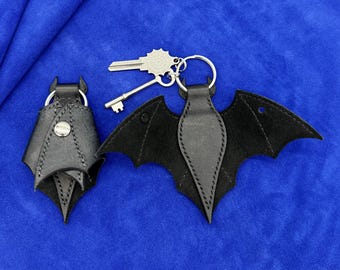 May include: Two black leather bat-shaped key holders. One is folded and secured with a silver metal snap button, while the other is unfolded to show its wing shape. A silver metal key ring with two attached keys is connected to the unfolded bat.