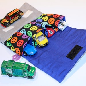 May include: A blue and gray fabric toy car carrier with a velcro closure. The carrier has three pockets with smiley faces printed on them. There are five toy cars inside the pockets, including a red, yellow, blue, white, and black car. A green toy truck is sitting in front of the carrier.