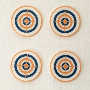 May include: Four circular coasters with a target design. Each coaster has concentric circles in white, navy blue, and orange, with a bullseye at the centre. The coasters are arranged in a square formation against a white background.