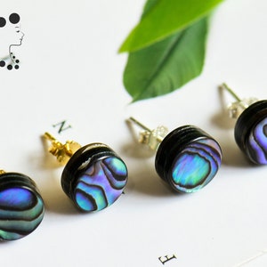 May include: Four stud earrings featuring iridescent abalone shells set in black metal. The earrings have a post backing. The earrings are displayed on a white surface with a logo of a woman's silhouette.