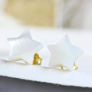 May include: A pair of star-shaped stud earrings. The earrings are white with a pearlescent sheen, set on gold-colored posts. The earrings are displayed on a white surface, with a soft, blurred background.