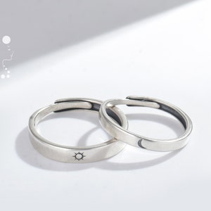 Könnte beinhalten: Zwei verstellbare Silberringe. Ein Ring zeigt ein Sonnendesign, der andere einen Mond. Die Ringe werden auf einer weißen Oberfläche präsentiert. Die Ringe sind wahrscheinlich aus Sterlingsilber.