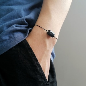 May include: A black bracelet featuring a rectangular, dark-colored stone. The stone is strung on a thin, black cord. The bracelet is worn on a wrist, with a blue shirt and black pants in the background.