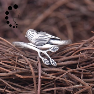 Puede incluir: Un anillo plateado con un pájaro detallado posado en una rama con hojas. El pájaro tiene plumas texturizadas y un ojo pequeño. El diseño del anillo envuelve el dedo, creando un accesorio inspirado en la naturaleza.