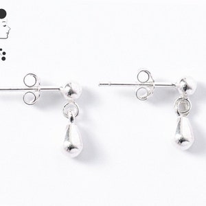 Puede incluir: Un par de pendientes de plata con un pequeño perno redondo y un colgante en forma de lágrima. Los pendientes se muestran sobre un fondo blanco. Diseño sencillo y elegante.