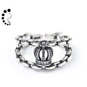 May include: A silver-toned open ring featuring a double chain band design connected by a central crown motif. The crown is detailed with small clear stones along the base and vertical ridges, while the chain links have a polished finish.