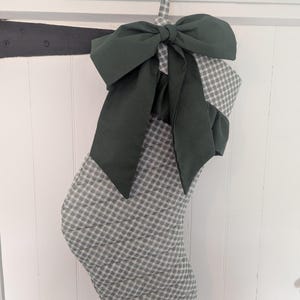 May include: A quilted Christmas stocking featuring a grey and white gingham check pattern. The stocking is adorned with a large, solid dark forest green fabric bow at the top, which sits above a matching dark green ruffled fabric trim detail.