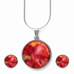 Könnte beinhalten: Ein Schmuckset mit einer Anhängerkette und passenden Ohrsteckern. Der Anhänger und die Ohrringe haben einen runden, silberfarbenen Rahmen. Das Design im Inneren des Rahmens zeigt eine lebendige Mischung aus roten, rosa und goldenen Farben.