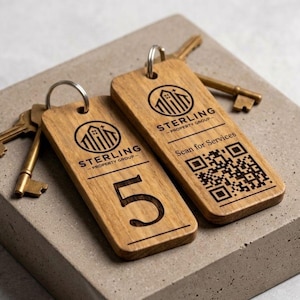 May include: Two wooden keychains with metal rings and brass keys. The keychains have the "STERLING - PROPERTY GROUP" logo and the number "5" and a QR code. The keychains are on a gray concrete surface.
