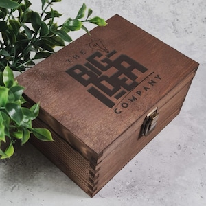 May include: A wooden box with a dark brown finish. The box features the text "THE BIG IDEA COMPANY" in a bold, stylized font. A metal clasp secures the box. Green foliage is visible in the background.