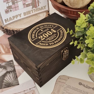 May include: A dark brown wooden box with a hinged lid and metal clasp. The box has a circular gold emblem on the top that reads "VINTAGE 2004 ORIGINAL PARTS AGED TO PERFECTION." A birthday card with the words "HAPPY BIRTHDAY" is in the background.