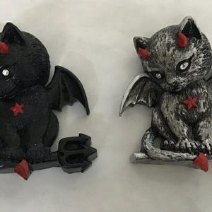 May include: Two cat figurines with bat wings, horns, and a trident. One is solid black, and the other is silver-grey with black highlights. Both feature red horns, a red star on the chest, a red tail tip, and small clear rhinestones for eyes.