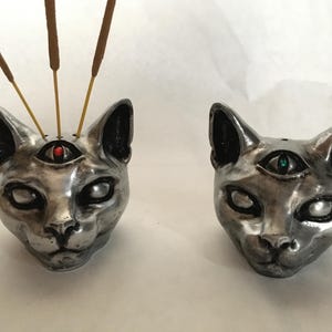May include: Two silver cat head incense holders. One holds incense sticks, the other is empty. Each cat head has a third eye with a gemstone. One has a red gemstone, the other has a green gemstone. The cat heads are detailed and metallic.