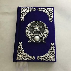 May include: A rectangular journal covered in deep purple velvet fabric. The front cover features four silver-toned ornate corner filigree pieces with embedded clear crystals and a central silver emblem depicting a pentagram within a crescent moon.