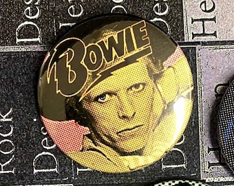 Puede incluir: Un botón redondo con una imagen en negro y amarillo de David Bowie. El botón tiene la palabra "BOWIE" en letras negras grandes en la parte superior.