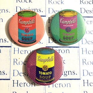 Puede incluir: Tres botones redondos con un diseño pop art que presenta una lata de sopa Campbell's. Las latas son amarillas, rojas y azules con un fondo blanco. El texto en las latas dice "Campbell's Condensed Tomato Soup".