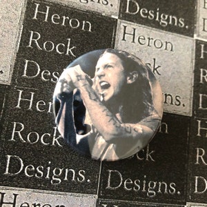 May include: A black and white button featuring a close-up image of a person singing into a microphone. The person has long hair and is wearing a dark shirt.