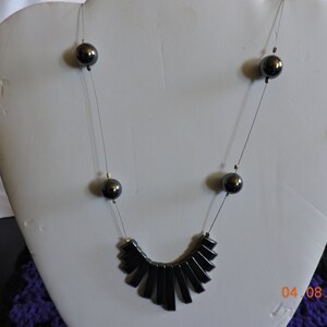 Puede incluir: Un collar negro con un colgante en forma de abanico hecho de cuentas rectangulares negras. El collar está enhebrado en una fina cadena plateada y tiene pequeñas cuentas redondas negras espaciadas a lo largo de la cadena.