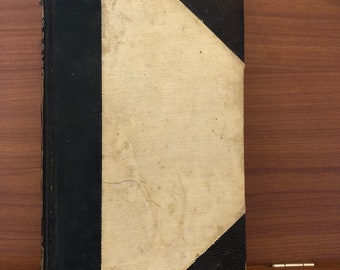 May include: A vintage book with a black leather spine and a cream-colored cloth cover. The cover has a worn and faded appearance.
