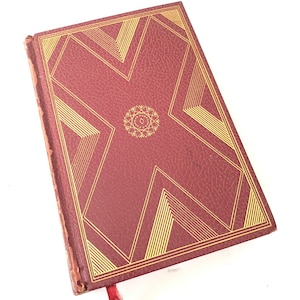 May include: A burgundy-colored book with gold geometric designs on the cover. The cover features a central circular design and a red ribbon bookmark. The book's edges show signs of wear, suggesting it is vintage or well-loved.