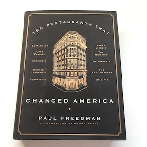 May include: A black book cover with gold lettering that reads "Ten Restaurants That Changed America" by Paul Freedman with an introduction by Danny Meyer. The cover features a gold illustration of a building with windows and a storefront.