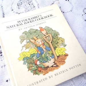May include: A vintage cookbook titled "Peter Rabbit's Natural Foods Cookbook" by Arnold Dobrin, illustrated by Beatrix Potter. The cover features a cartoon illustration of Peter Rabbit holding a carrot and a robin perched on a garden tool.
