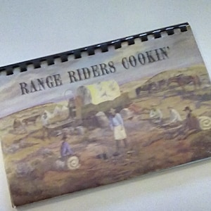 May include: A vintage cookbook titled "Range Riders Cookin'" with a colorful illustration of a group of people gathered around a campfire.