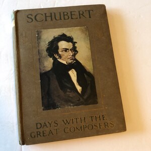 May include: A vintage book titled "SCHUBERT" with a portrait of a man in glasses. The book cover is brown with the text "DAYS WITH THE GREAT COMPOSERS" in embossed lettering. The book is slightly worn.
